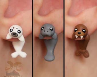 May include: Three pairs of polymer clay earrings shaped like seals. The first pair is white, the second pair is gray, and the third pair is brown.