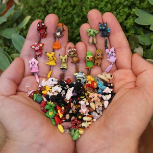 May include: A collection of colorful, handmade polymer clay earrings featuring various cartoon characters, animals, and abstract designs. The earrings are displayed on a person's hand.