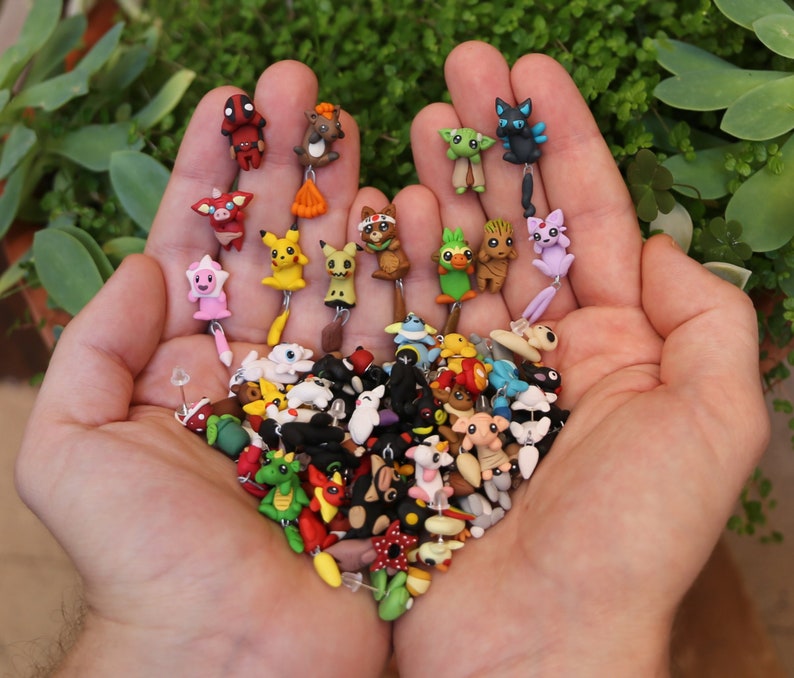 May include: A collection of colorful, handmade polymer clay earrings featuring various cartoon characters, animals, and abstract designs. The earrings are displayed on a person's hand.