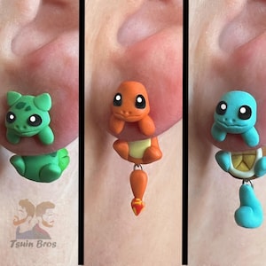 Könnte beinhalten: Drei handgefertigte Ohrringe aus Polymer Clay mit beliebten Zeichentrickfiguren. Die Ohrringe sind in Form eines grünen, schildkrötenartigen Wesens, eines orangefarbenen, echsenartigen Wesens und eines blauen, schildkrötenartigen Wesens.