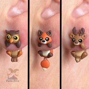 Könnte beinhalten: Drei Paar ausgefallene Ohrringe mit Waldtier-Designs. Ein Paar zeigt eine braune Eule mit gelben Augen, ein anderes einen Fuchs mit orangefarbenem Schwanz und das letzte ein Reh mit Geweih. Die Ohrringe sind am Ohr einer Person befestigt.