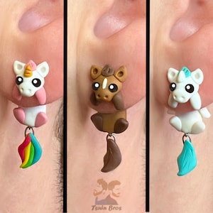 Könnte beinhalten: Drei Ohrstecker mit modellierten Tierfiguren: ein weißes Einhorn mit goldenem Horn und Regenbogen-Schweif, ein braunes Pferd mit dunkelbraunem Schweif und ein weißes Einhorn mit türkisfarbenem Horn und Schweif. Jeder Ohrring hat ein hängendes Element.