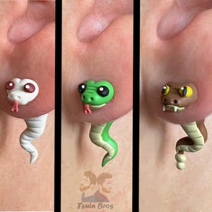 May include: Three pairs of snake-shaped earrings in white, green, and brown. Each earring features a snake head and a curved body. The snakes have red tongues and are worn on an ear. The earrings are handmade.