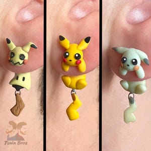 May include: Three handmade polymer clay earrings featuring Pokémon characters. One earring depicts Mimikyu, another Pikachu, and the third a green Pokémon. Each earring has a dangling charm. The earrings are attached to a person's ear.