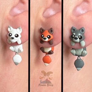 Può includere: Tre paia di orecchini originali con disegni di animali. Un paio è bianco con una coda bianca, un altro è arancione con una coda abbinata e il terzo è grigio con una coda grigia. Gli orecchini hanno un ciondolo a forma di coda pendente.