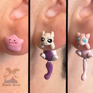 Op de afbeelding: Een set van drie handgemaakte oorstekers met Pokémon-personages. Eén is een roze Ditto, en de andere twee zijn Mewtwo en Mew. De oorbellen zijn bevestigd aan het oor van een persoon. De merknaam "Tsuin Bros" is zichtbaar.