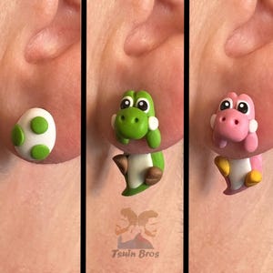 Può includere: Un set di tre orecchini: un uovo bianco e verde, un Yoshi verde e un Yoshi rosa. Gli orecchini sono progettati per sembrare che escano dall'orecchio. Il testo "Tsuin Bros" è visibile.