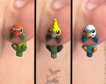 May include: Three pairs of colorful bird-shaped earrings. One pair features a green and orange bird, another a yellow and gray bird, and the last a blue and white bird. The earrings are attached to a person's ear. The brand name "Tsuin Bros" is visible.