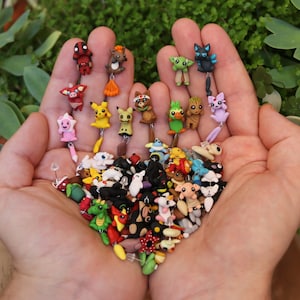May include: A collection of colorful, handmade polymer clay earrings featuring various cartoon characters, animals, and abstract designs. The earrings are displayed on a person's hand.