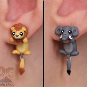 May include: A pair of dangle earrings featuring a lion and an elephant. The lion is brown with a yellow mane and the elephant is gray with white tusks. Both animals have black eyes and are made of polymer clay.