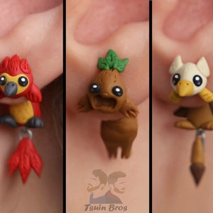 May include: Three pairs of handmade polymer clay earrings. The earrings feature colorful, cartoonish designs of a bird, a plant-like creature, and a bird-like creature. Each earring has a dangling element. The earrings are attached to a person's ear.