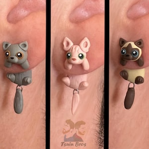 May include: Three pairs of novelty earrings with cute animal designs. Each earring set features a different animal: a grey puppy, a pink Sphynx cat, and a Siamese cat. Each earring has a dangling tail charm. The Tsuiin Bros logo is visible.