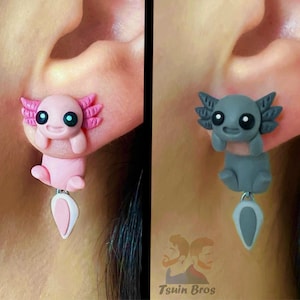 May include: A pair of dangle earrings featuring a pink and a gray axolotl charm. The axolotl charms are made of polymer clay and have a cute, cartoon-like appearance. The earrings are hanging from a silver hook.