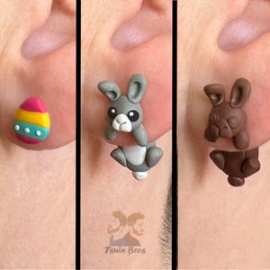 May include: Close-up of three Easter-themed earrings. One is a colourful egg with pink, yellow, and blue stripes. The other two are bunny-shaped, one grey and white, the other brown. The earrings are handmade.