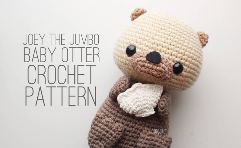 K&ouml;nnte beinhalten: Ein geh&auml;keltes Stofftier-Otter namens Joey, das ein wei&szlig;es geh&auml;keltes Herz h&auml;lt. Das Spielzeug ist braun und cremefarben mit schwarzen Augen und einer schwarzen Nase. Der Text "JOEY THE JUMBO BABY OTTER CROCHET PATTERN" ist in schwarzer Schrift auf wei&szlig;em Hintergrund geschrieben.