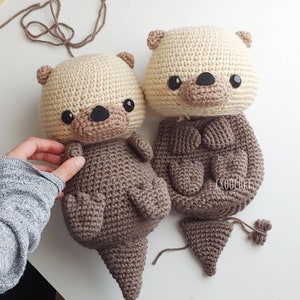K&ouml;nnte beinhalten: Zwei geh&auml;kelte Otter-Pl&uuml;schtiere, eines braun und eines beige, mit schwarzen Augen und Nasen. Der braune Otter ist komplett, w&auml;hrend dem beige Otter der Schwanz fehlt und ein separates Schwanzst&uuml;ck vorhanden ist.