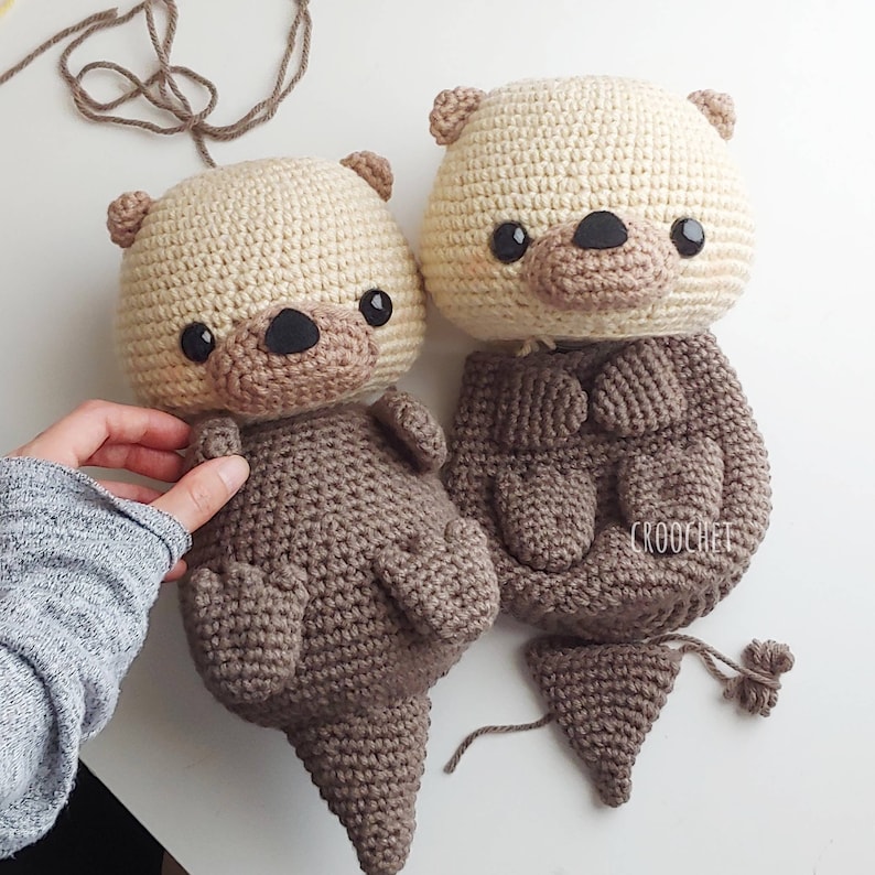 K&ouml;nnte beinhalten: Zwei geh&auml;kelte Otter-Pl&uuml;schtiere, eines braun und eines beige, mit schwarzen Augen und Nasen. Der braune Otter ist komplett, w&auml;hrend dem beige Otter der Schwanz fehlt und ein separates Schwanzst&uuml;ck vorhanden ist.
