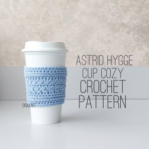 Peut inclure: Un manchon de tasse en crochet bleu clair sur un gobelet à café blanc à emporter. Le texte "Astrid Hygge Cup Cozy Crochet Pattern" est imprimé en noir sur un fond gris.