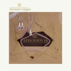 May include: A silver jewelry set featuring teardrop-shaped earrings and a matching pendant necklace. The earrings hang from silver hooks, and the necklace has a delicate chain. The jewelry is displayed on a textured, tan background with the brand name "PATHOS JOYAS CA" visible.