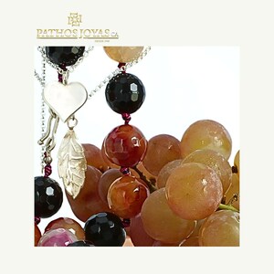 May include: A close-up of a necklace with a silver heart and leaf charm, and black, red, and orange gemstone beads. The necklace is displayed next to a bunch of grapes. The logo "PATHOS JOYAS CA DESDE 1996" is in the top left corner.