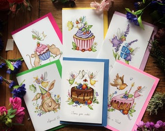 Op de afbeelding: Een verzameling wenskaarten met grillige illustraties. Elke kaart heeft een ander ontwerp, waaronder cupcakes, taarten, feeën en een konijn. De kaarten hebben kleurrijke randen en zijn omgeven door bloemen.