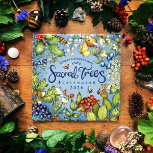 Op de afbeelding: Een kalender voor 2026 getiteld "Sacred Trees" met een blauwe omslag met illustraties van vlinders, bessen en feeën. De kalender is omgeven door natuurlijke elementen zoals bladeren, dennenappels en bessen op een houten oppervlak.