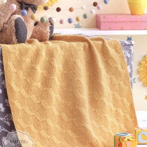 Puede incluir: Una manta de punto amarillo dorado con un patrón geométrico está sobre un estante. Un oso de peluche con pajarita está detrás de la manta. La imagen también incluye una guirnalda de pompones de colores, una caja rosa y bloques de juguete.