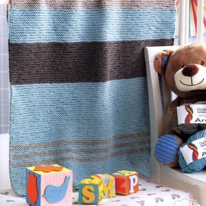 May include: A knitted baby blanket in shades of blue, brown, and gray, with a striped pattern. The blanket is draped over a white crib. Beside the blanket are colorful toy blocks spelling out "SMP" and a stuffed teddy bear.