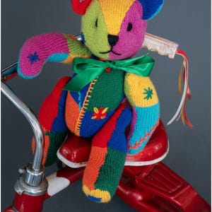 Puede incluir: Un oso de peluche tejido multicolor con un diseño de parches en hilo rojo, verde, amarillo, azul y morado, con detalles de estrellas bordadas y una cinta de satén verde atada al cuello. El texto PDF File PATTERN es visible.