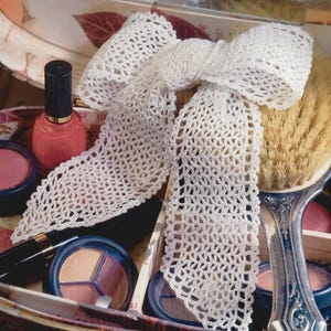 Pode incluir: Um laço branco de crochê de trama aberta está sobre uma maleta de maquiagem vintage cheia de produtos de beleza. Um frasco de esmalte coral, sombra para os olhos e um pincel de maquiagem são visíveis. O texto "PDF File PATTERN" está impresso no canto inferior direito.