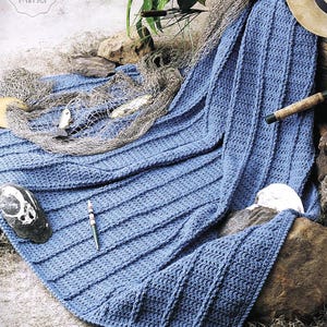 May include: A blue crochet blanket with a ribbed design is draped over rocks. A fishing net with decorative fish, a pen, and a seashell are placed near the blanket. The image also includes a straw hat and greenery.