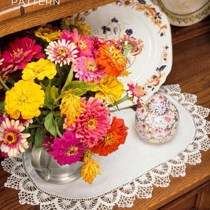 May include: A vibrant bouquet of zinnias in a silver vase, with blooms in shades of yellow, pink, and orange. The arrangement sits on a white lace doily, next to a floral-patterned ceramic jar and a decorative plate. The text "PDF File Pattern" is visible.