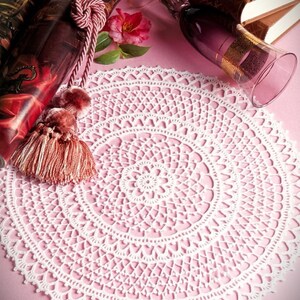 May include: A round white crochet doily featuring an intricate, repeating geometric lace pattern with a central floral motif and a scalloped edge. The doily is displayed on a pink surface alongside a dark red tassel and a purple glass goblet.