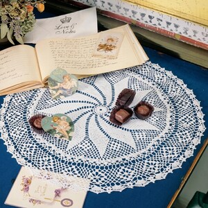 May include: A white, crocheted doily with a spiral design is the centerpiece of this image. The doily is surrounded by vintage ephemera, including a heart-shaped card with an angel illustration, a handwritten letter, and chocolates.