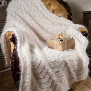 May include: A white knitted throw blanket with a textured chevron pattern and a fuzzy, mohair-like appearance. The blanket is draped over a chair and features a small, square, light brown gift box tied with a gold ribbon on its surface.