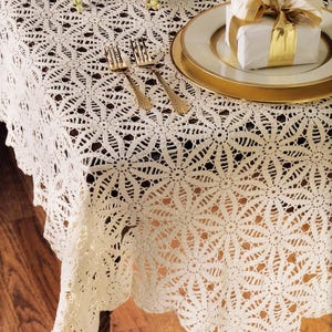 May include: Cream-colored, crocheted tablecloth with a floral pattern. Two gold forks and a gold-rimmed plate with a wrapped gift are on the table. The tablecloth has a scalloped edge and is draped over a wooden surface.