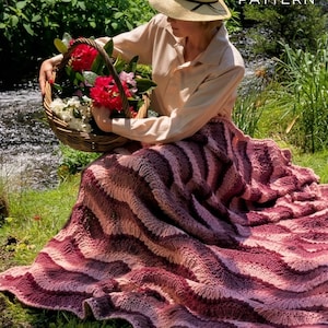 Puede incluir: Una persona con sombrero de paja y camisa clara está sentada en la hierba cerca de un arroyo, sosteniendo una cesta de mimbre llena de flores rojas y blancas. Una gran manta tejida ondulada en tonos rosa y burdeos está extendida sobre la hierba. El texto en la imagen dice "PDF File PATTERN".