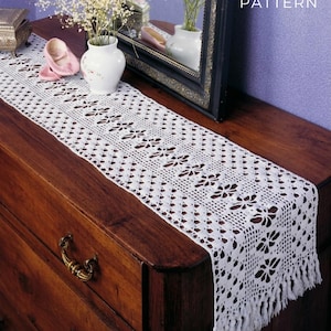 May include: A white, open-weave crochet table runner with a floral pattern and fringe. The runner is displayed on a dark wood dresser. The image also includes a mirror, vase, and decorative items.