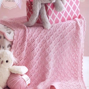 Puede incluir: Una manta de bebé de punto rosa claro con un patrón de diamantes y bordes festoneados. Un conejito de peluche gris y una caja con estampado rosa y blanco están encima de la manta. Un búho de peluche blanco y un ovillo de lana rosa están al lado. El texto "Knitting PDF Pattern" es visible.