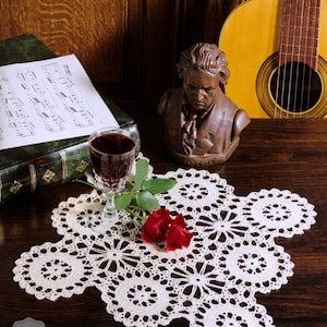 Może przedstawiać: Kremowa, szydełkowa serwetka z kwiatowym wzorem, kieliszek czerwonego wina i czerwona róża. Popiersie Beethovena i gitara klasyczna w tle. Serwetka leży na ciemnej drewnianej powierzchni. Tekst "CROCHET PDF PATTERN" znajduje się w lewym dolnym rogu.