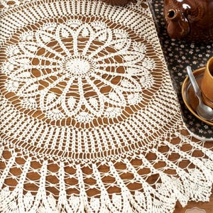 Puede incluir: Un tapete redondo de ganchillo de color crema con un intrincado diseño floral. El tapete está colocado sobre una superficie de madera, con una tetera y una taza marrones en el fondo. El detallado patrón del tapete añade un toque de elegancia a cualquier mesa.