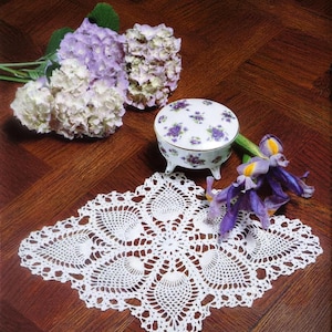 May include: A white crocheted doily with a complex geometric pattern, a small round porcelain trinket box featuring a purple floral print and gold trim, and several purple and white hydrangea and iris flowers. Text reads PDF File PATTERN.
