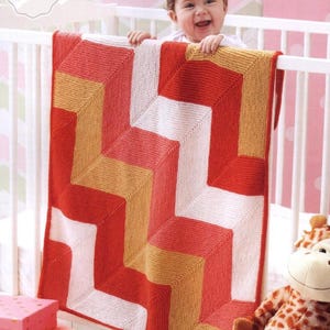 May include: A knitted baby blanket with a geometric pattern in red, white, and gold hangs over a crib. The blanket features a zig-zag design. The words "Knitting PDF Pattern" are in the upper left corner. A baby smiles behind the crib.