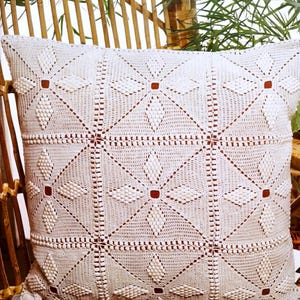 May include: A white, square crochet cushion with a geometric floral pattern. The cushion features a repeating design of diamond shapes and flower-like motifs, with small red squares at the centre of each flower. The cushion is resting on a wicker chair.