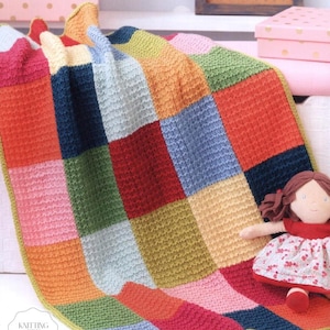 May include: A colorful, patchwork knitted blanket with squares of red, orange, blue, green, and yellow. A small doll with a red dress and brown hair rests on the blanket. The blanket has a textured appearance and a green border.