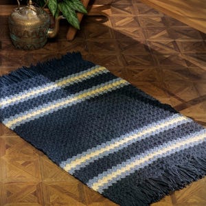 May include: A rectangular, hand-crocheted rug with a navy blue base and horizontal stripes of light blue and yellow. The rug has a fringed edge and is displayed on a wooden floor. The words "PDF File Pattern" are visible in the lower left corner.