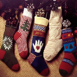 Puede incluir: Cinco calcetines de Navidad tejidos con varios diseños, incluyendo copos de nieve, rayas y una mano con un corazón. Los calcetines son en tonos de rojo, verde, azul y crema, con lazos decorativos. El texto "Christmas Stockings" es visible.