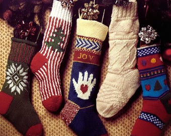 May include: A collection of five knit Christmas stockings with various designs, including snowflakes, stripes, and a hand with a heart. The stockings are in shades of red, green, blue, and cream, with decorative bows at the top. The text "Christmas Stockings" is visible.