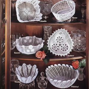 May include: A display of white crochet bowls in various designs, including square, oval, and round shapes. The text "Cut Glass Bowls" and "Crochet" are visible at the top. The bowls are displayed on a wooden shelf.