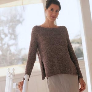 May include: A woman wearing a brown sweater and a light gray skirt. The sweater has a boat neck and long sleeves. The skirt is knee-length. The woman is leaning against a white rocking chair. The image also includes the text "KNITTING PDF PATTERN".